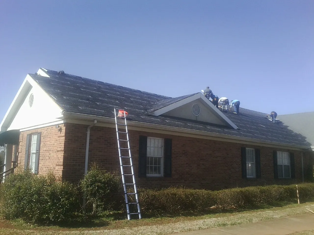 Crew tearing shingles off a brick ranch roof with a ladder set — Emergency Roof Repair work by our crew in Cogan Station