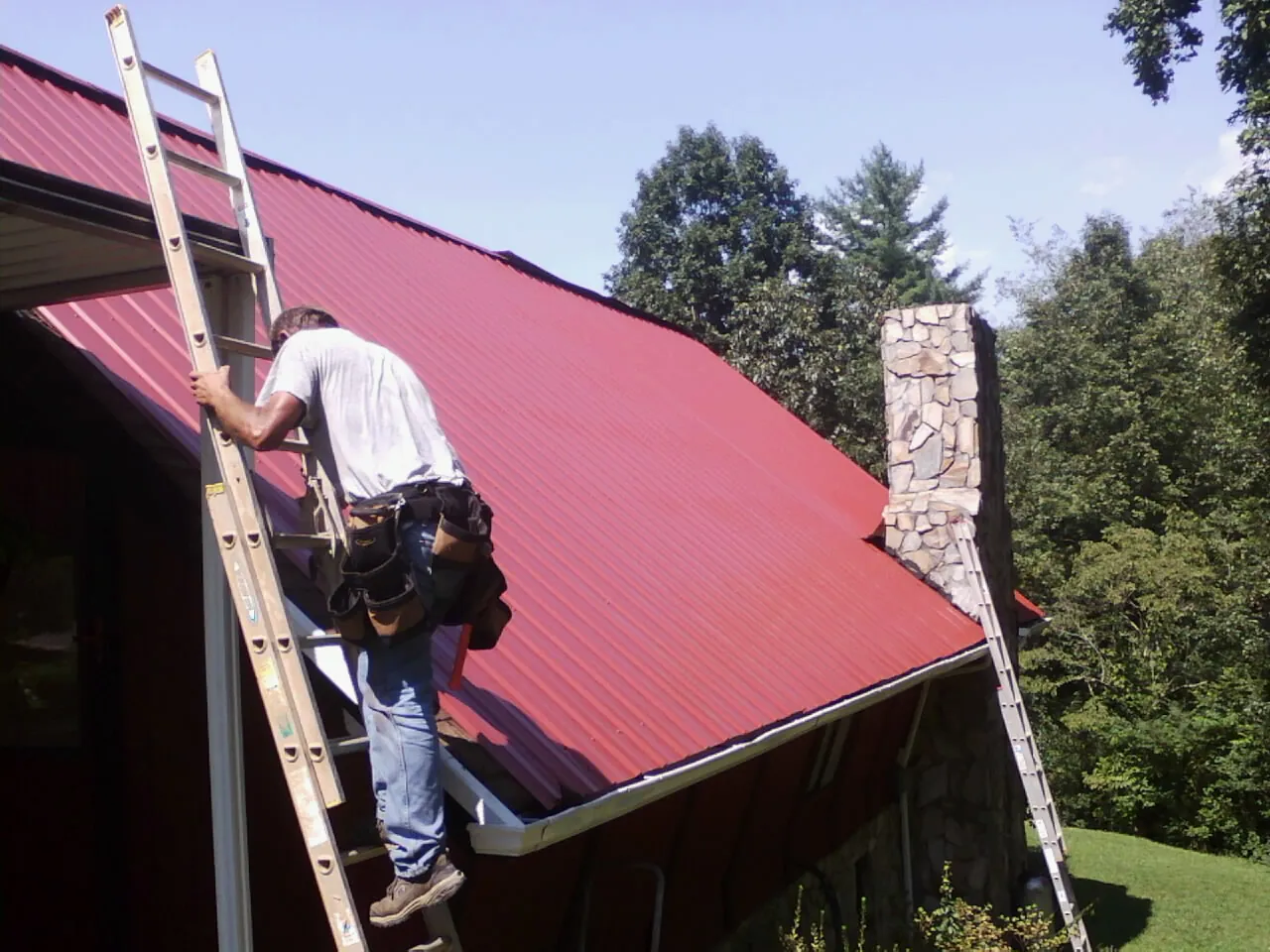 Roofer on a ladder beside a red metal roof and stone chimney — Soffit & Fascia Repair by Roofing Brothers in Cogan Station
